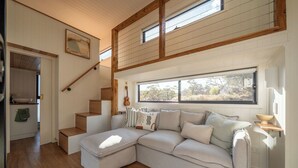 Un interior moderno con un amplio sofá seccional, piso de madera, una escalera y un ventanal con vistas a árboles.