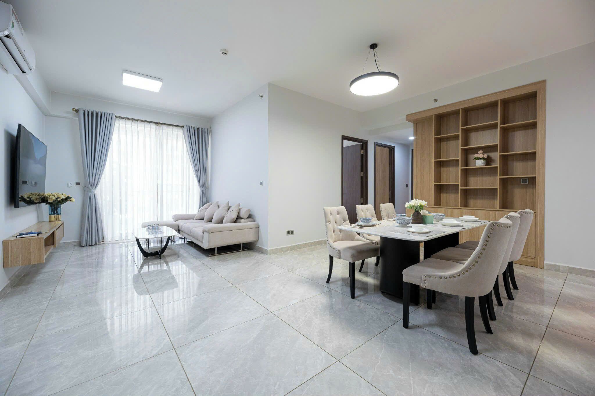Een moderne woonkamer met een bank, een eettafel en stoelen. Er is een televisie aan de muur bevestigd en een groot raam met gordijnen.