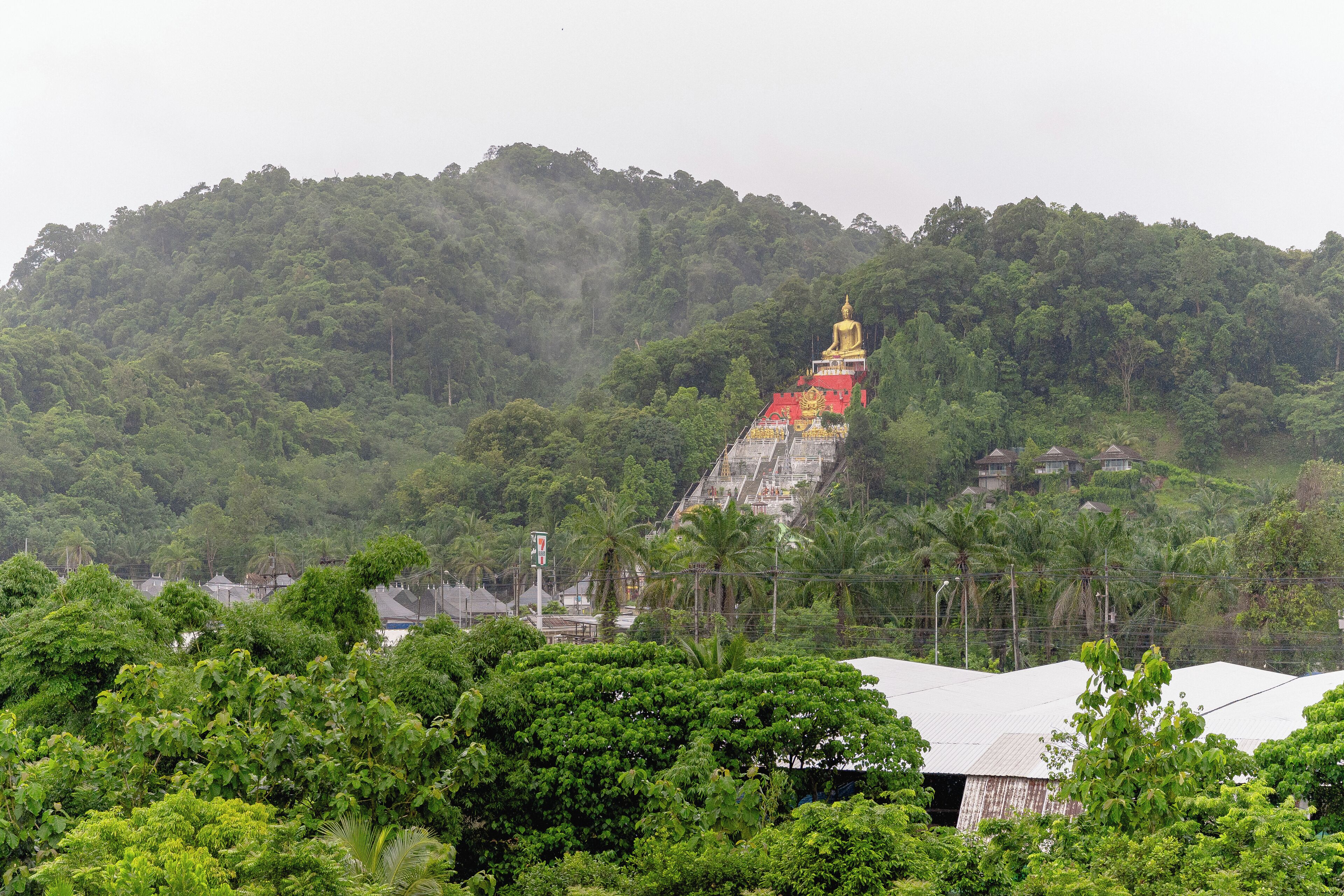 A temple with a golden statue on a hill, surrounded by lush greenery and residential buildings.