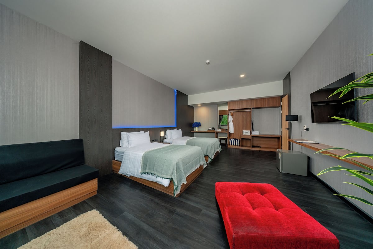 A modern hotel room with two beds, a sofa, a red ottoman, and a TV on the wall.