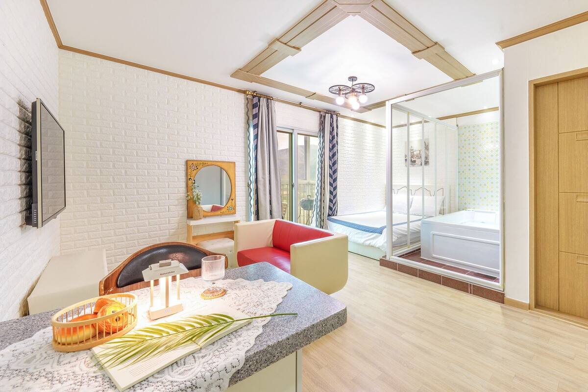 Une chambre d’hôtel moderne avec un grand lit, un coin salon avec un fauteuil rouge, une petite table avec un vase et une plante, et une grande fenêtre avec des rideaux.