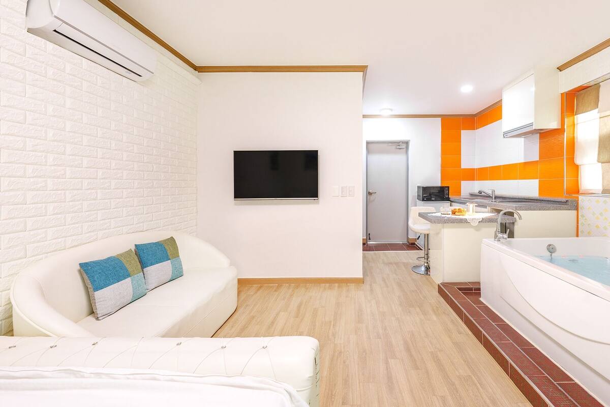 A modern bathroom with a bathtub, a sink, and a toilet. There is a TV mounted on the wall and a window providing natural light.