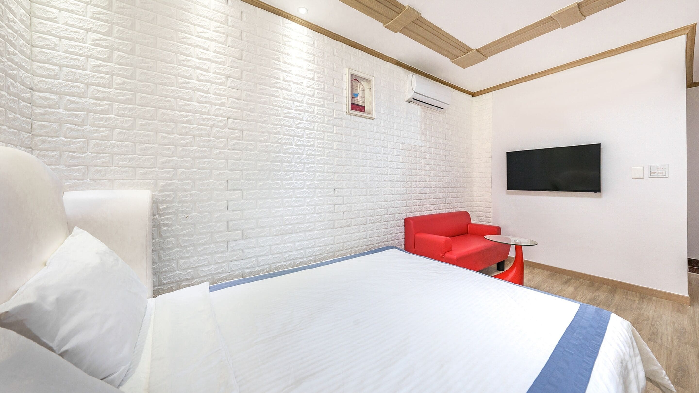 A hotel room with a white brick wall, a bed with white bedding, a red chair, a small glass table, and a flat-screen TV mounted on the wall.