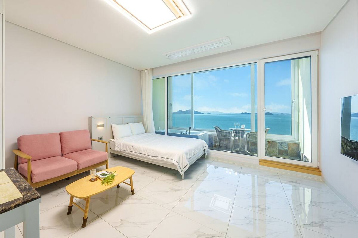 Ett modernt hotellrum med en säng, en rosa soffa, ett litet träbord och ett stort fönster med utsikt över havet.