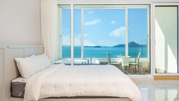 A hotel room with a large bed, a view of the sea, and a balcony with a table and chairs. - Yeosu Stay Raonpine Spa Pension (Yeosu)