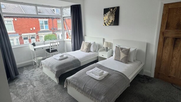 A twin bedroom with two beds, a desk, and a window with a view of a red brick building. - Simonside Apartment - Sleeps 6 - Pets Ok - Parking (Newcastle upon Tyne)