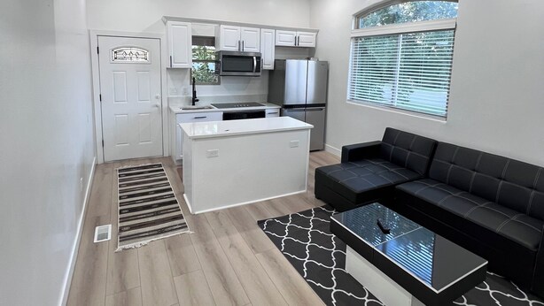 A modern kitchen with white cabinets, a built-in microwave, and a refrigerator. There is a black leather sofa and a glass coffee table in the living area.