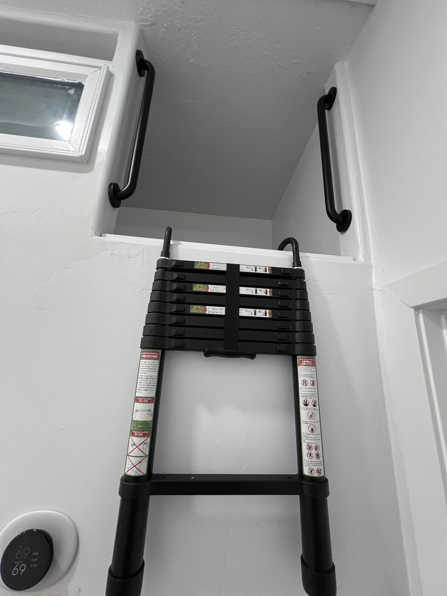 Een zwarte metalen ladder met veiligheidsinstructies, bevestigd aan een witte muur met een raam erboven.