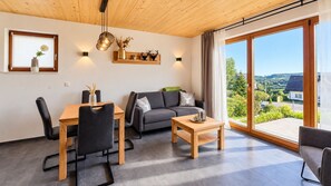 A modern living room with a dining area, a sofa, a coffee table, and a view of a green landscape through large windows.