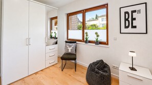 A room with a bed, a chair, a window with blinds, a bedside table, and a wall-mounted lamp.