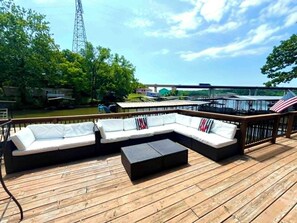 Eine Holzterrasse mit einer Eckcouch und einem kleinen Tisch, mit Blick auf ein Gewässer, im Hintergrund eine Brücke und viel Grün.