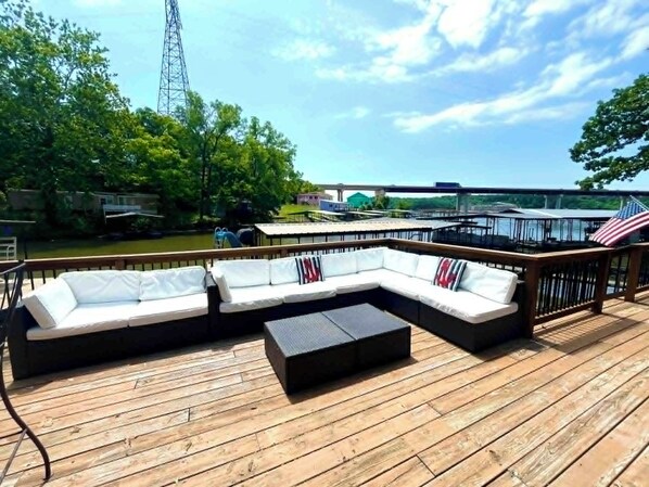 A wooden deck with a sectional sofa and a small table, overlooking a body of water with a bridge and greenery in the background.