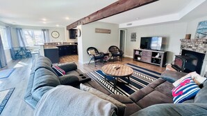Un salon moderne avec un grand canapé, une table basse et une cheminée. On aperçoit, au fond, un coin cuisine avec une table à manger et des chaises.