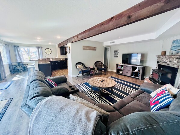 A modern living room with a large sofa, a coffee table, and a fireplace. There is a kitchen area in the background with a dining table and chairs.
