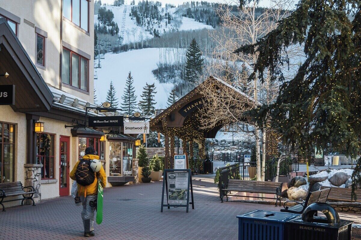 Paisaje montañoso cubierto de nieve con una estructura de madera decorada con luces, un camino pavimentado, bancos y un contenedor de reciclaje.