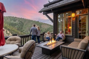 Une terrasse avec coin salon extérieur et foyer, offrant une vue sur une colline boisée.