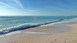 Ein Strand mit sanft anschwellenden Wellen, wolkenlosem Himmel und einer fernen Küstenlinie.