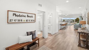 A spacious living area with a wooden bench, a framed sign reading "Madeira Beach 27° 47' 53.11" N 82° 47' 50.48" W, and a view of the kitchen and dining area.