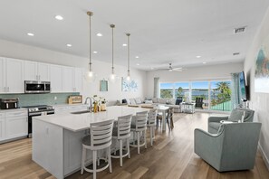 A modern kitchen with white cabinetry, a central island with bar stools, and a dining area with a view of the outdoors. - Luxury Beach Penthouse at Madeira Del Mar (Madeira Beach)