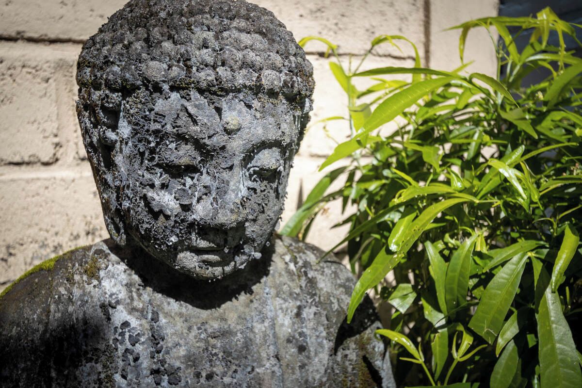 A stone statue with a rough texture, placed outdoors next to a brick wall and green foliage.