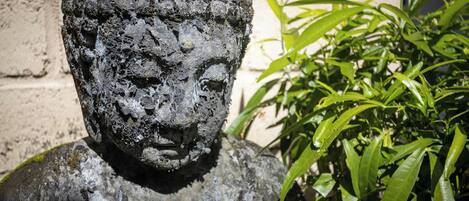 A stone statue with a rough texture, placed outdoors next to a brick wall and green foliage.