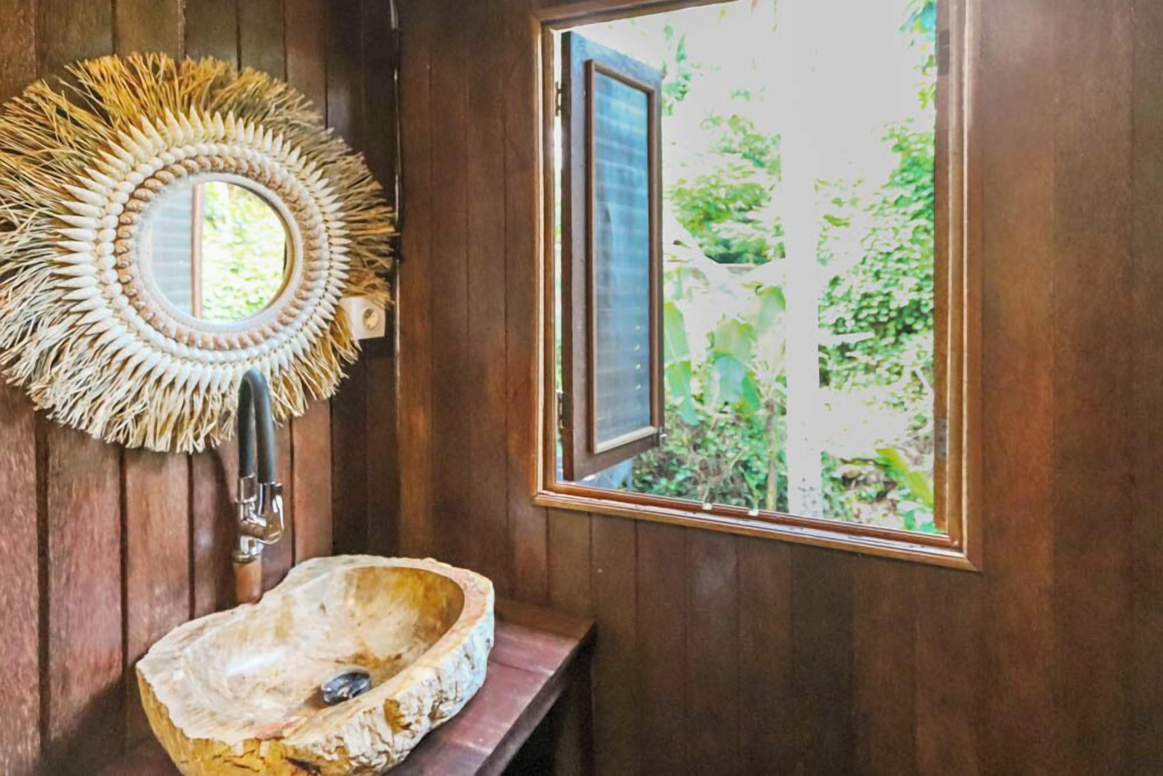 A rustic bathroom with a wooden sink, a woven mirror, and a window with a view of greenery.