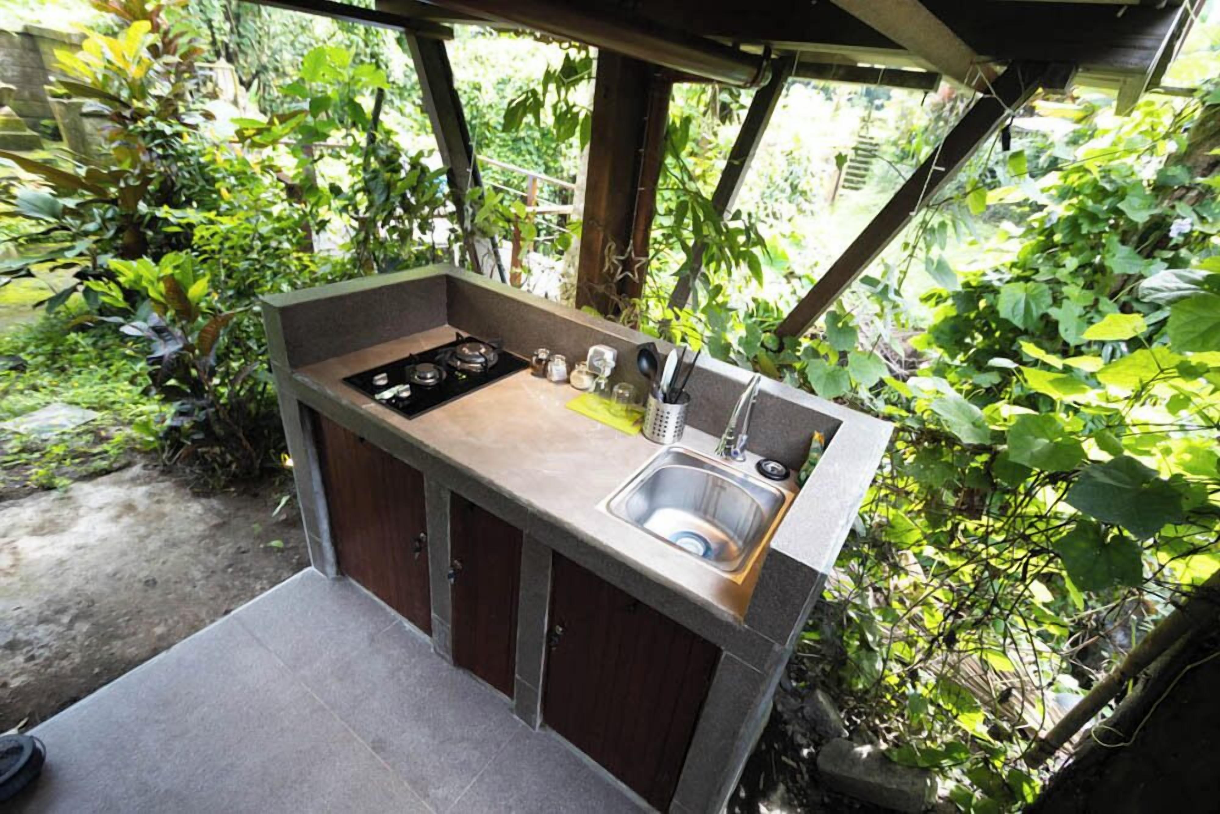 Une petite cuisine extérieure dotée d’une cuisinière à gaz, d’un évier et de placards en bois, le tout entouré d’une végétation luxuriante.