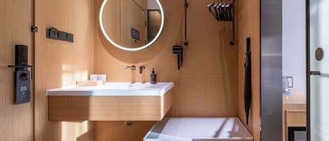 A modern bathroom with a wooden vanity, a round mirror, a towel rack, and a towel dispenser.