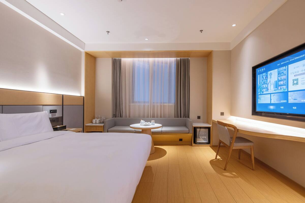 A modern hotel room with a large bed, a desk, a sofa, and a TV screen displaying a menu.