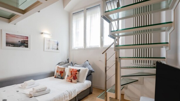 Ein Schlafzimmer mit einem Bett, Kissen, einem Nachttisch, einem Fenster und einer Glaskonstruktion als Treppe.