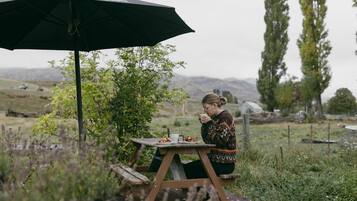 Une personne est assise à une table en bois, tenant un parapluie noir. La scène se déroule à l’extérieur, dans un espace verdoyant, avec des arbres et des collines en arrière-plan.