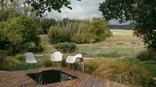 Una terraza de madera con tres sillas blancas, un pequeño estanque y vistas a un campo cubierto de césped y árboles.