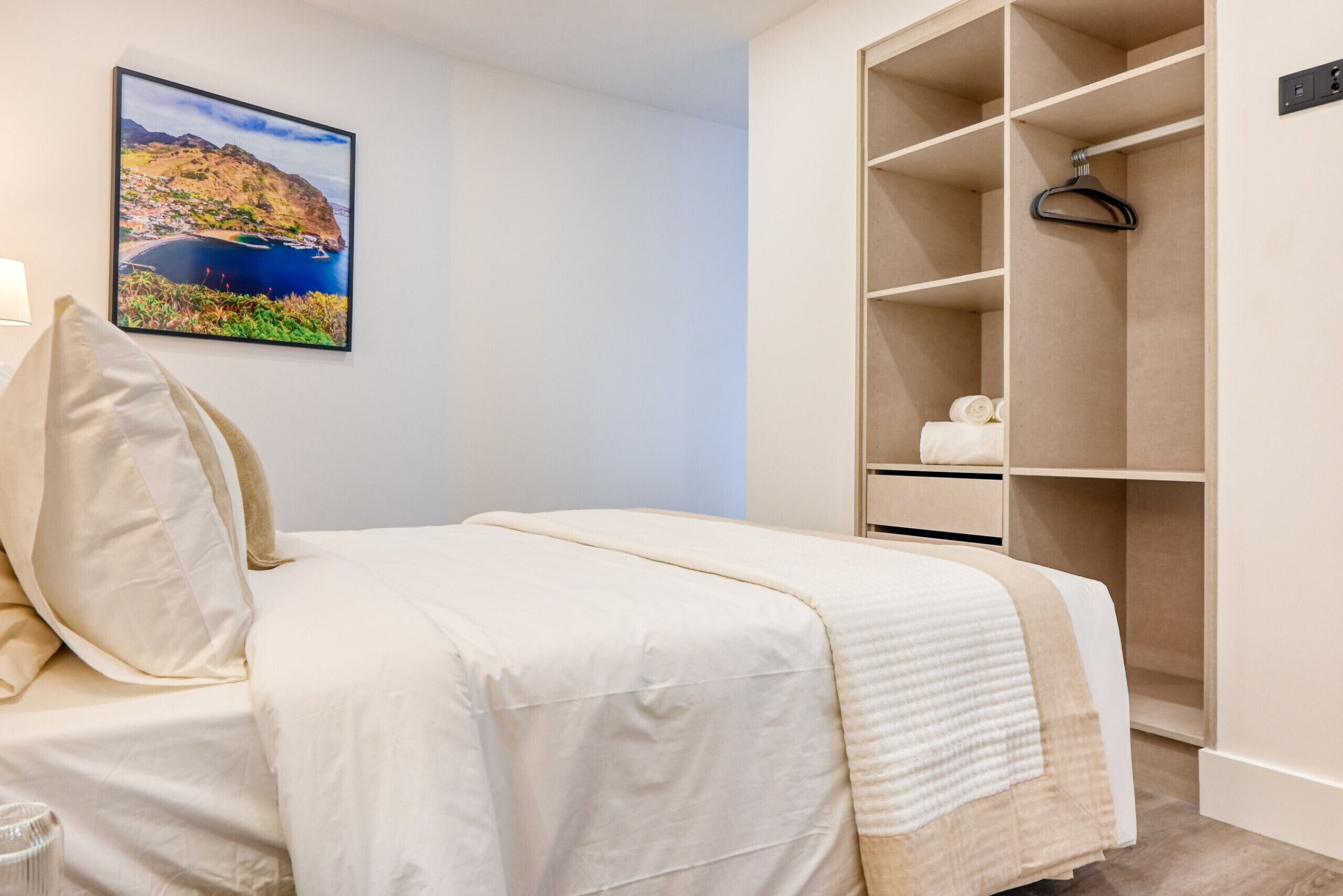 A bedroom with a bed, a wardrobe, a hanging clothes rack, and a framed picture on the wall.