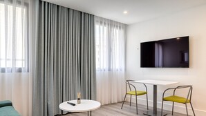 Une chambre d’hôtel moderne avec une grande fenêtre, des rideaux gris, une table blanche avec deux chaises jaunes, un canapé vert et un téléviseur à écran plat fixé au mur.