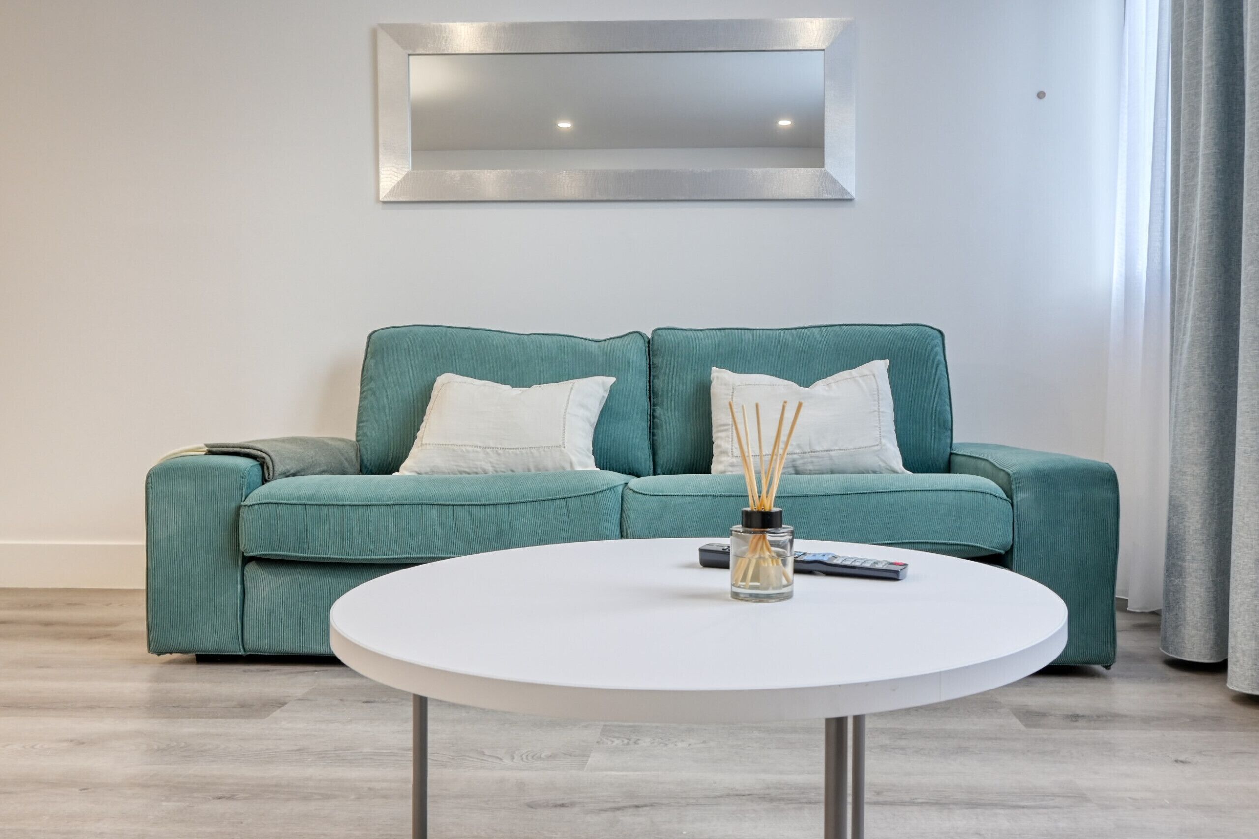 Un sofá color turquesa con cojines blancos, una mesa redonda blanca con un control remoto y una botella de vidrio, y un espejo moderno en la pared.