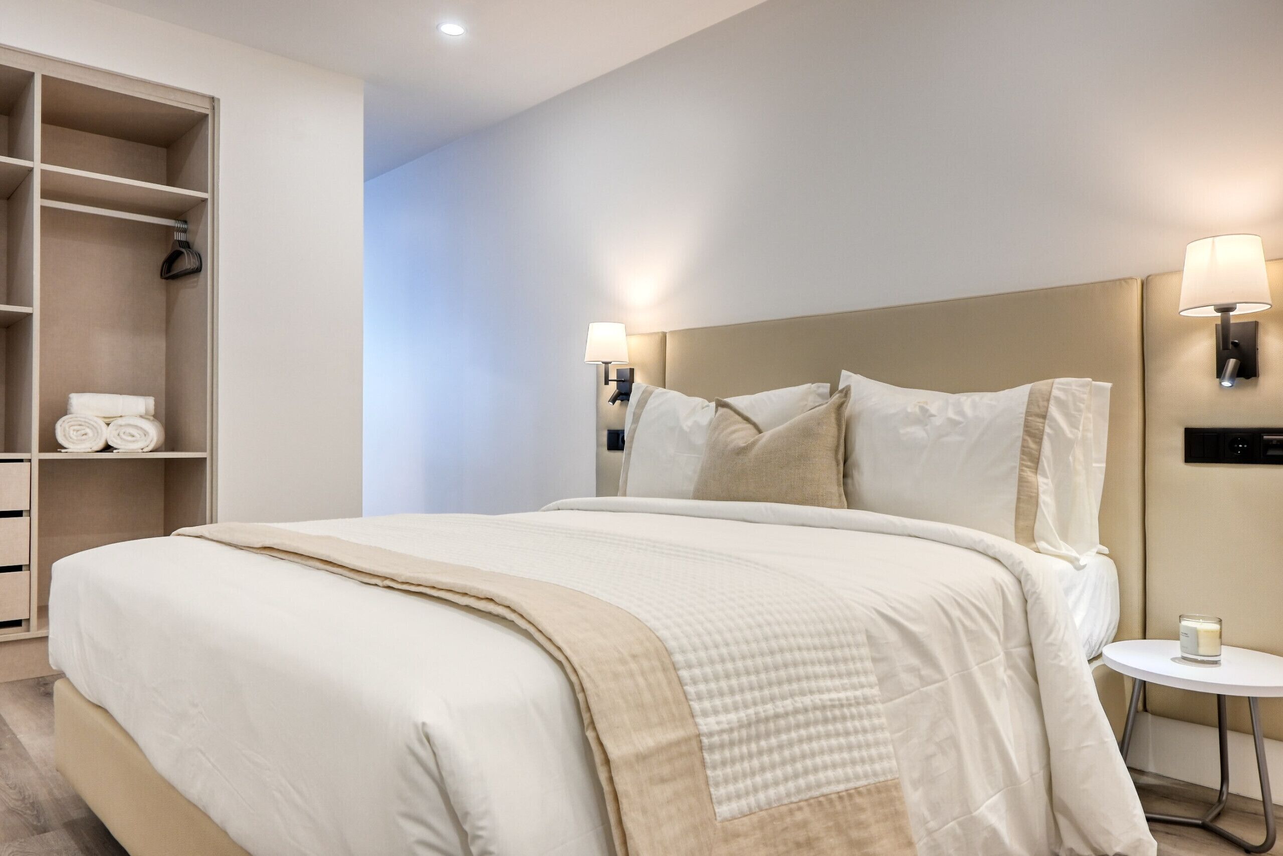 A neatly made bed with a beige headboard and pillows, a bedside table with a lamp, and a wardrobe with folded towels.