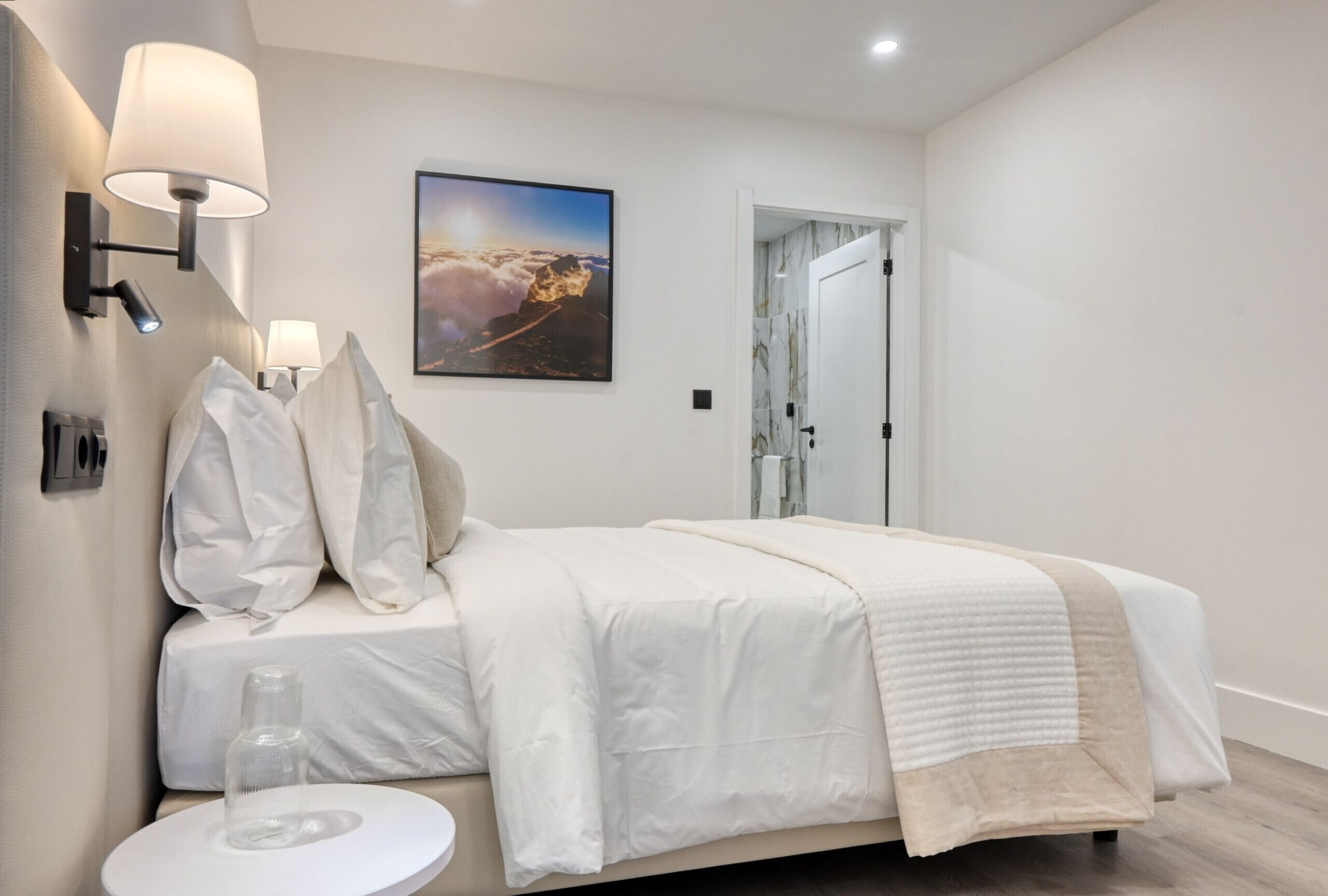 Une chambre d’hôtel moderne avec un lit soigneusement fait, une petite table ronde sur laquelle se trouve une bouteille en verre, et un tableau représentant un paysage de montagne accroché au mur.