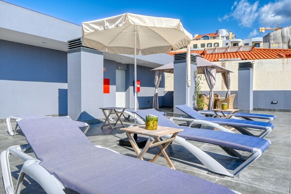 A rooftop lounge area with sun loungers, a table with a drink, and umbrellas.