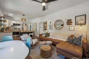 Un soggiorno moderno con un divano marrone, poltrone blu, un tavolo bianco e una cucina sullo sfondo.