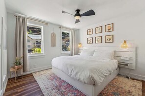 A bedroom with a bed, a ceiling fan, a window with curtains, a wall-mounted lamp, and framed pictures.