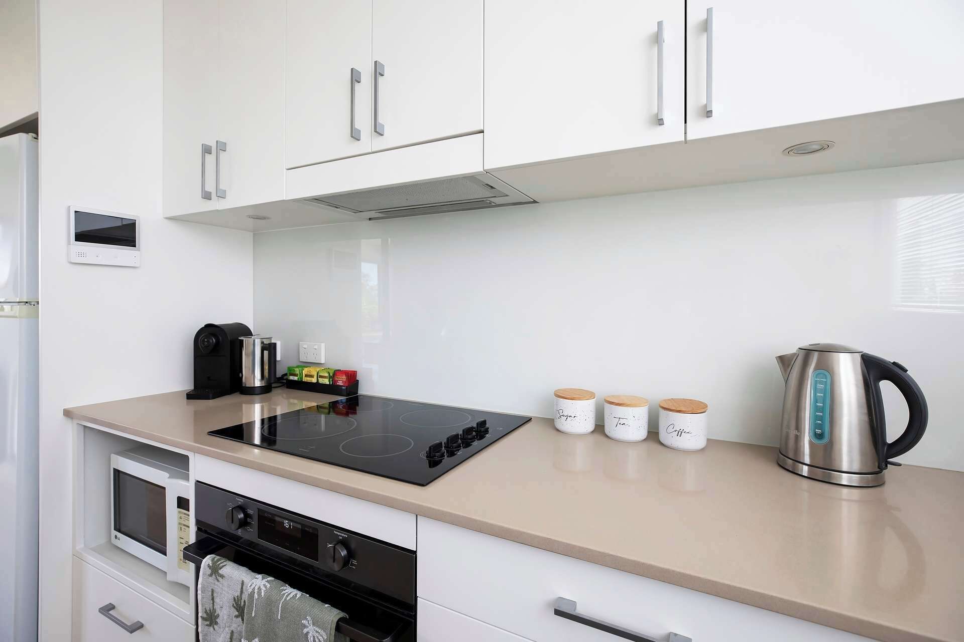 Eine moderne Küche mit weißen Schränken, einer schwarzen Induktionskochplatte, einem Edelstahl-Wasserkocher und drei weißen Gläsern mit Holzdeckeln.