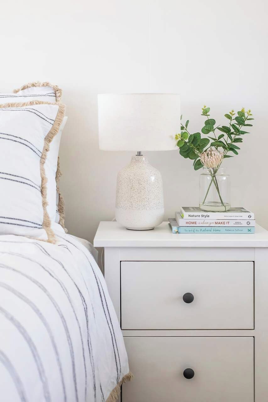 Een keurig opgemaakt slaapkamer met een bed, een nachtkastje met een lamp, een vaas met bloemen, en een stapel boeken.