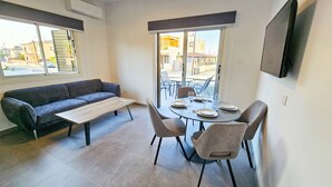 Un coin repas comprenant une table ronde, quatre chaises, un canapé gris et une fenêtre avec des stores.