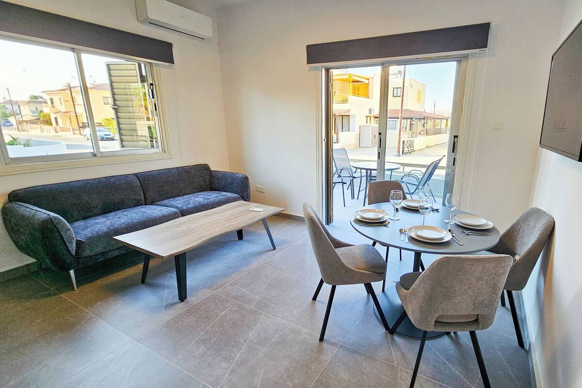 Un coin repas comprenant une table ronde, quatre chaises, un canapé gris et une fenêtre avec des stores.