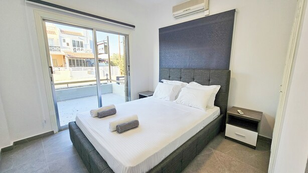 Une chambre moderne avec un grand lit, une table de chevet, une fenêtre donnant sur des immeubles et un appareil de climatisation.