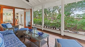 A spacious room with a glass table, wicker furniture, and large windows offering a view of greenery.