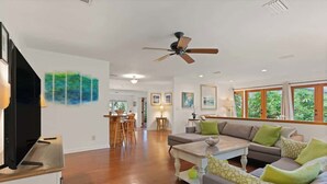 A spacious living area with a sofa, coffee table, and a TV on a stand. There is a ceiling fan, artwork on the walls, and a window with a view of greenery.