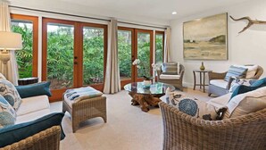 A spacious living room with wicker furniture, a glass coffee table, and large windows offering a view of lush greenery.