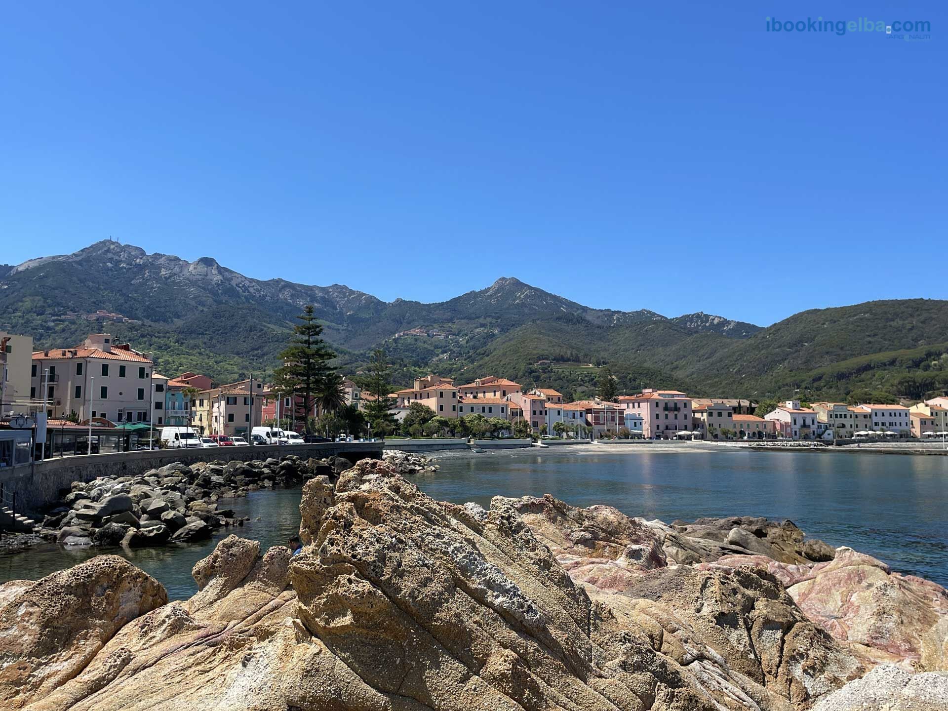 A coastal town with buildings along the waterfront, rocky terrain in the foreground, and mountains in the background.