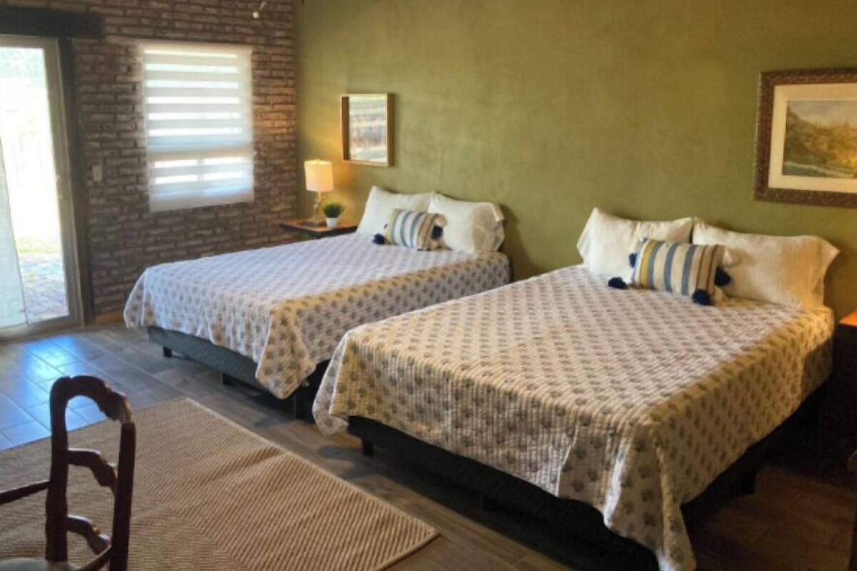 Quarto com duas camas, parede de tijolos, janela com persianas, luminária e um quadro na parede.